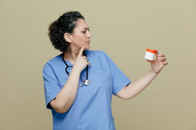 Nachdenkliche Ärztin mittleren Alters in Uniform und Stethoskop um den Hals, die das Kinn berührt und den Tablettenbehälter isoliert auf olivfarbenem Hintergrund betrachtet