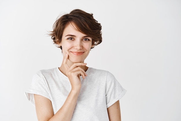 Nachdenkliche schöne Frau mit kurzen, unordentlichen Haaren, lächelt und berührt saubere perfekte Haut ohne Make-up, stehend auf weißer Wand