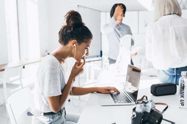 Nachdenkliche schwarze Frau, die in den Kopfhörern nahe Laptop sitzt, während ihre Kollegen neben Flipchart stehen. Innenporträt der Fotografin, die mit Computer arbeitet und auf Tastatur im Büro tippt.