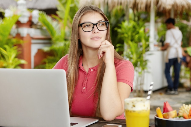 Nachdenkliche selbständige Frau mit Brille, die vor einem offenen Laptop sitzt, sich auf den Ellbogen stützt und wegschaut, während sie in den Ferien aus der Ferne arbeitet.