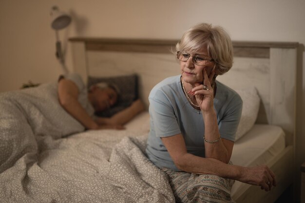 Nachdenkliche Seniorin, die auf dem Bett sitzt und sich Sorgen um etwas macht Ihr Mann schläft im Hintergrund