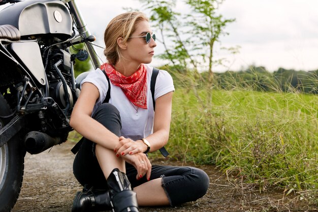 Nachdenkliche, sorglose, entspannte Bikerin trägt stilvolle Sonnenbrillen, ein weißes T-Shirt und Jeans, sitzt auf Asphalt in der Nähe eines Motorrads und ist tief in Gedanken versunken. Junge Frau schaut in die Ferne, ruht sich nach der Fahrt aus
