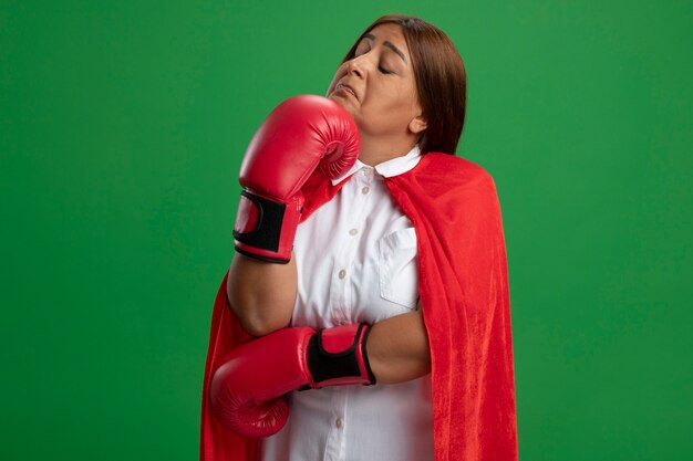 nachdenkliche Superheldenfrau mittleren Alters mit geschlossenen Augen, die Boxhandschuhe tragen, die Hand unter Kinn lokalisiert auf Grün setzen