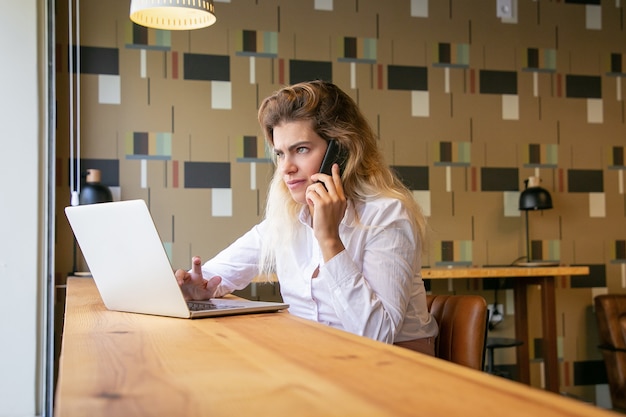 Nachdenkliche Unternehmerin, die Laptop verwendet und auf Mobiltelefon im gemeinsamen Arbeitsraum spricht