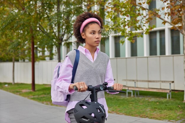 Nachdenkliche weibliche Fahrten Elektroroller pendelt mit Öko-Transport zur Schule oder Universität trägt Rucksack-Posen auf der Straße im Freien konzentriert in die Ferne. Jugend- und urbanes Lifestyle-Konzept