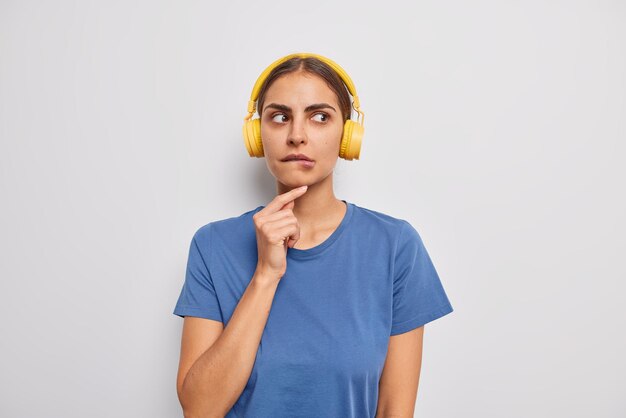 Nachdenkliche, zweifelnde Frau beißt Lippenkonzentrate weg, hat nachdenklichen Ausdruck, hört Audiospur über drahtlose Kopfhörer, trägt lässiges blaues T-Shirt, das über weißem Hintergrund isoliert ist. Lass mich nachdenken