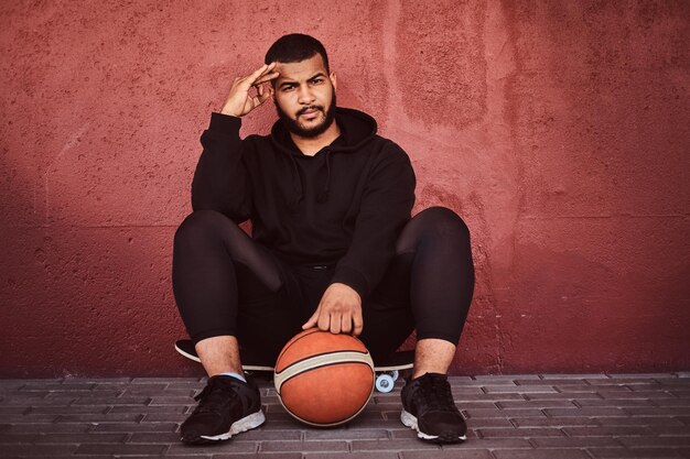 Nachdenklicher afroamerikanischer bärtiger Typ in schwarzem Hoodie und Sportshorts, der auf einem Skateboard mit Basketball sitzt und sich an die Wand lehnt.