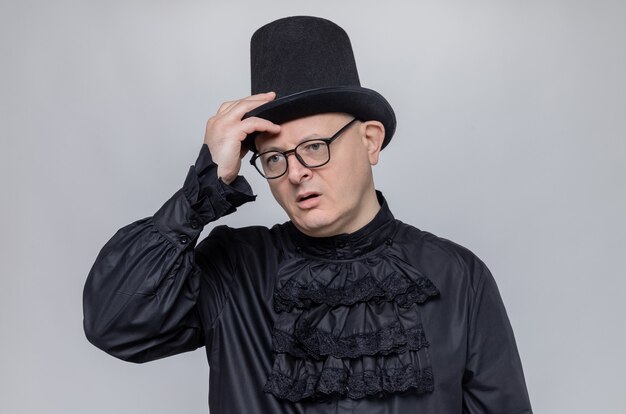 Nachdenklicher erwachsener Mann mit Hut und Brille in schwarzem Gothic-Hemd, der die Hand auf seinen Hut legt und zur Seite schaut
