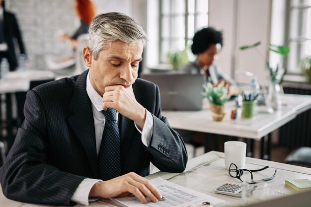 Nachdenklicher Geschäftsmann, der am Schreibtisch im Büro sitzt und Finanzberichte durchgeht