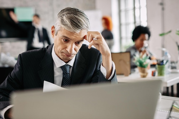 Kostenloses Foto nachdenklicher geschäftsmann, der darüber nachdenkt, wie er probleme lösen kann, während er im büro papierkram erledigt es gibt menschen im hintergrund