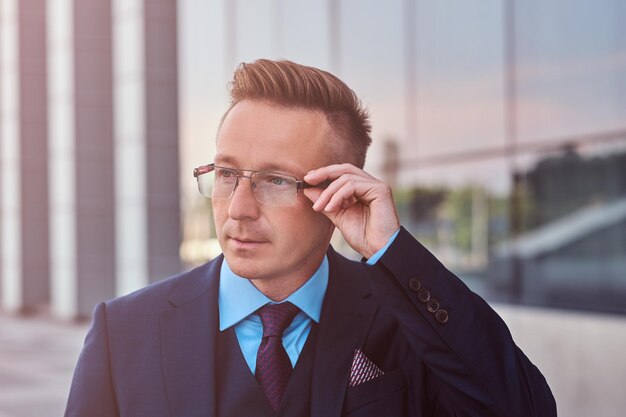 Nachdenklicher Geschäftsmann in einem eleganten Anzug, der wegschaut und seine Brille korrigiert, während er im Freien vor Wolkenkratzerhintergrund steht.