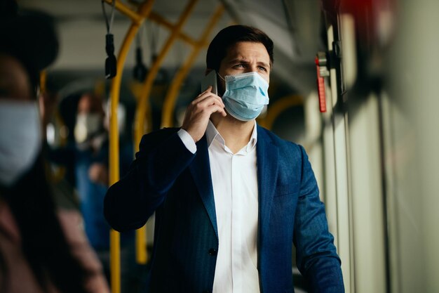 Nachdenklicher Geschäftsmann mit schützender Gesichtsmaske telefoniert, während er mit dem Bus zur Arbeit pendelt