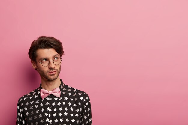 Nachdenklicher Hipster in modischem schwarzem Hemd und rosa Fliege, bereit für ein Date mit einer Freundin, überlegt, was er als Geschenk geben soll, trägt eine runde Brille