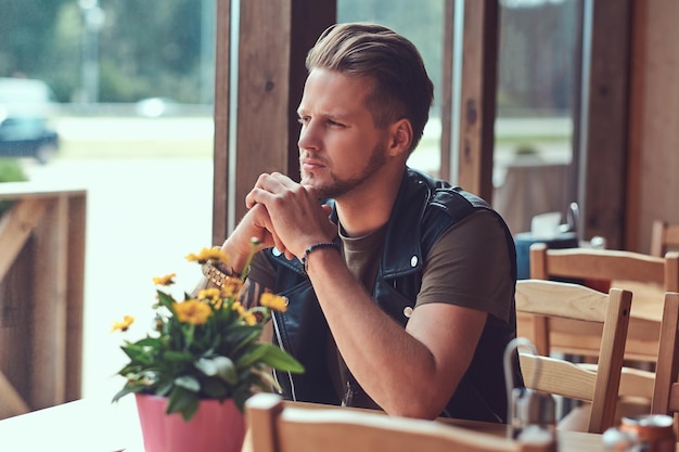 Nachdenklicher Hipster mit stylischem Haarschnitt und Bart sitzt an einem Tisch in einem Straßencafé und blickt auf die Straße.