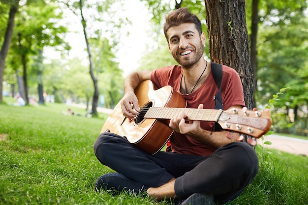 Nachdenklicher hübscher junger Mann, der Gitarre im Park spielt, sich auf Baum stützt und auf Gras sitzt