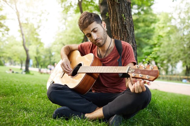 Nachdenklicher hübscher junger Mann, der Gitarre im Park spielt, sich auf Baum stützt und auf Gras sitzt