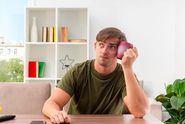 Nachdenklicher junger blonder gutaussehender Mann sitzt am Tisch und hält Tasse auf Gesicht und schaut zur Seite innerhalb des Wohnzimmers