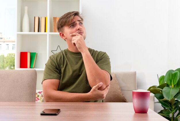 Nachdenklicher junger blonder hübscher Mann sitzt am Tisch mit Telefon und Tasse, die Kinn halten und im Wohnzimmer nach oben schauen