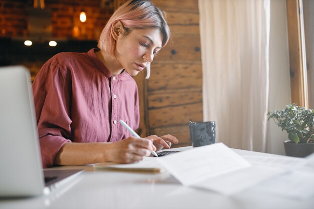 Nachdenklicher junger Büroangestellter, der Unterlagen überprüft, die von zu Hause aus arbeiten. Ernsthafte Frau, die Bericht liest, am Tisch mit Laptop sitzt.