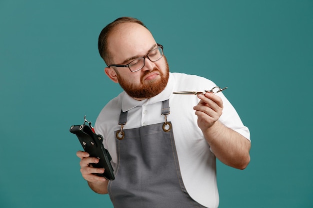 Nachdenklicher junger Friseur, der Uniform und Brille trägt und eine Friseurtasche mit Friseurwerkzeugen und Scheren hält, die eine Schere isoliert auf blauem Hintergrund betrachten