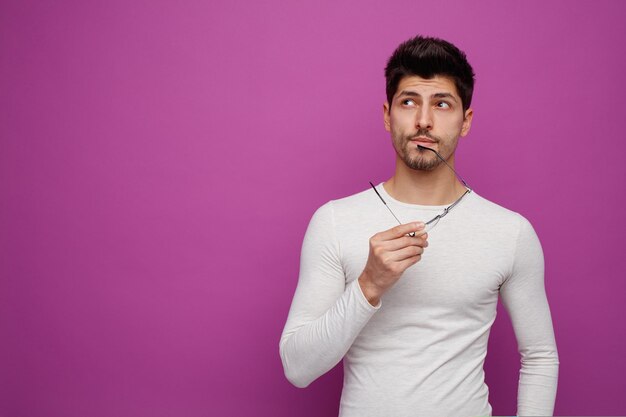 Nachdenklicher junger gutaussehender Mann, der eine Brille hält, die das Kinn berührt, mit einer Brille, die auf violettem Hintergrund mit Kopierraum zur Seite schaut