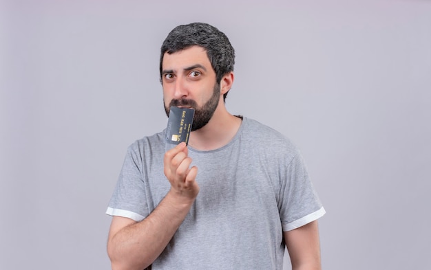 Nachdenklicher junger gutaussehender Mann, der Kreditkarte auf Mund lokalisiert auf weißer Wand setzt
