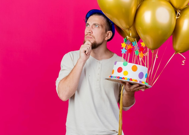 Nachdenklicher junger gutaussehender slawischer Party-Typ, der Partyhut hält, der Luftballons und Geburtstagstorte mit Sternen berührt, die Kinn berühren, das Seite lokalisiert auf rosa Wand mit Kopienraum betrachtet