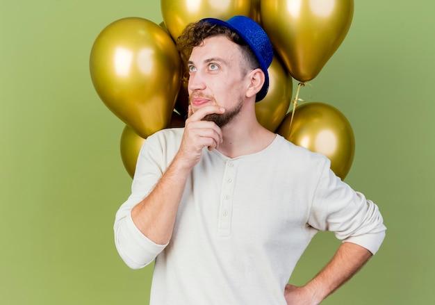 Nachdenklicher junger gutaussehender slawischer Party-Typ, der Partyhut trägt, der vor Luftballons steht und das berührende Kinn betrachtet, das Hand auf Taille hält, die auf olivgrüner Wand lokalisiert wird