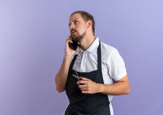 Nachdenklicher junger hübscher Friseur, der Uniform trägt, die am Telefon spricht, das oben schaut und Schere lokalisiert auf lila Wand hält