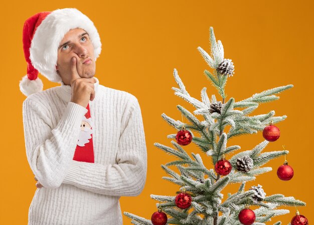 Nachdenklicher junger hübscher Kerl, der Weihnachtsmütze und Weihnachtsmannkrawatte trägt, die nahe verziertem Weihnachtsbaum steht, der Hand auf Kinn setzt, das lokalisiert auf orange Hintergrund schaut