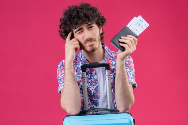 Nachdenklicher junger hübscher lockiger Reisender Mann, der Brieftasche und Flugtickets hält, die Arme auf Koffer und Finger auf Schläfe setzen, mit einem Auge geschlossen auf isolierter rosa Wand mit Kopienraum