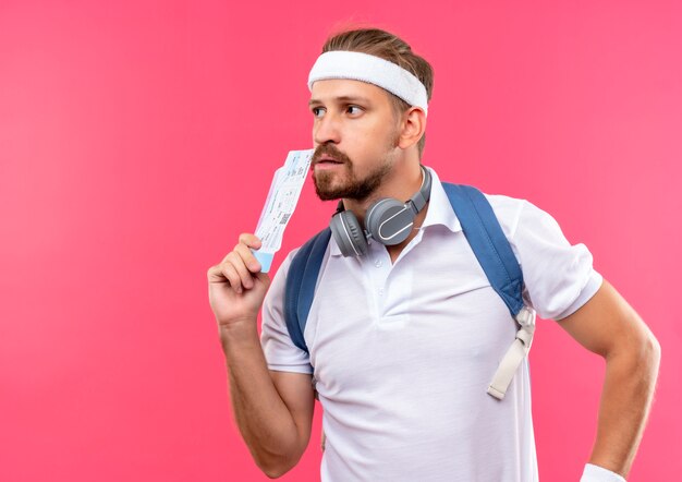 Nachdenklicher junger hübscher sportlicher Mann, der Stirnband und Armbänder und Rückentasche mit Kopfhörern am Hals trägt, die Flugtickets halten und Seite lokalisiert auf rosa Raum betrachten