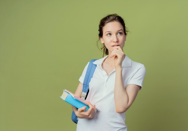 Nachdenklicher junger hübscher Student, der Rückentasche hält, die Buch und Notizblock hält, die Seite betrachtet und Hand nahe Mund lokalisiert auf olivgrünem Hintergrund mit Kopienraum hält