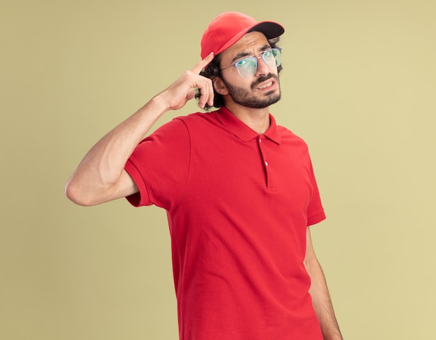 Nachdenklicher junger kaukasischer Liefermann in roter Uniform und Mütze mit Brille, der eine Denkgeste isoliert auf olivgrüner Wand macht