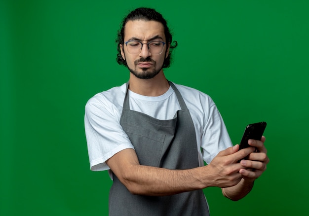 Nachdenklicher junger kaukasischer männlicher Friseur, der Uniform und Brille hält und Handy lokalisiert auf grünem Hintergrund mit Kopienraum hält