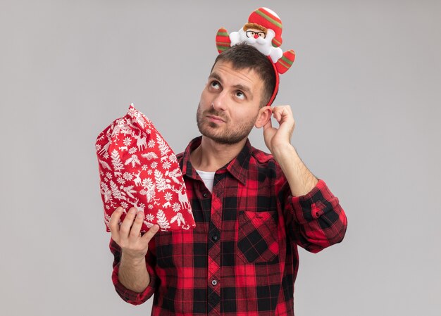 Nachdenklicher junger kaukasischer Mann, der Weihnachtsstirnband hält, das Weihnachtssack berührendes Ohr, das lokalisiert auf weißem Hintergrund hält