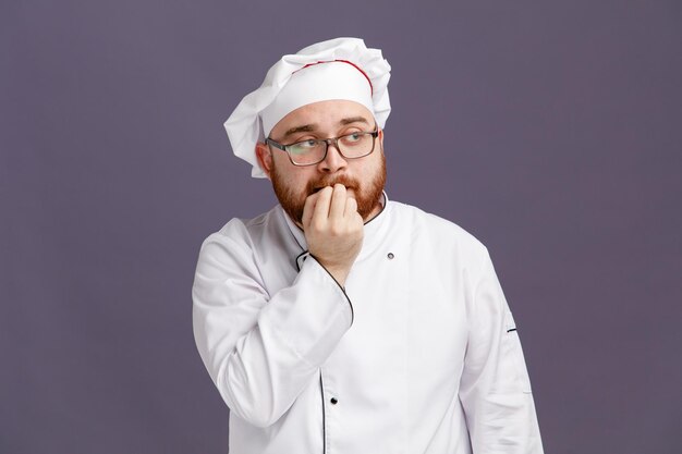 Nachdenklicher junger Koch mit Brille, Uniform und Mütze, der auf die beißenden Finger der Seite blickt, isoliert auf violettem Hintergrund