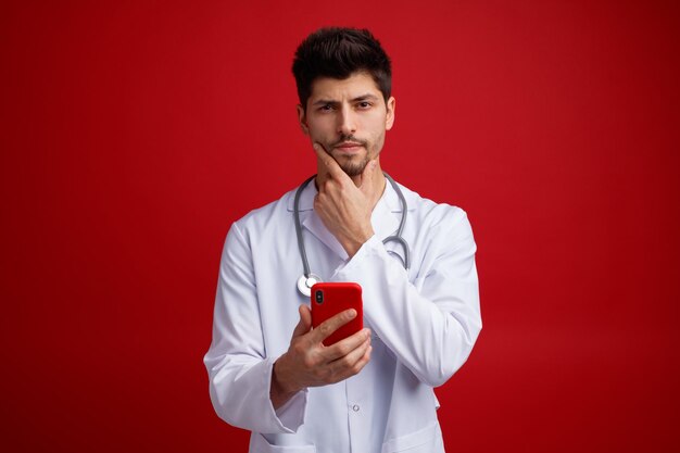 Nachdenklicher junger männlicher Arzt, der eine medizinische Uniform und ein Stethoskop um den Hals trägt und ein Handy hält, das die Hand am Kinn hält und isoliert auf rotem Hintergrund in die Kamera blickt