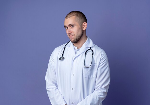 Nachdenklicher junger männlicher Arzt, der medizinisches Gewand und Stethoskop trägt, betrachtet Front lokalisiert auf lila Wand