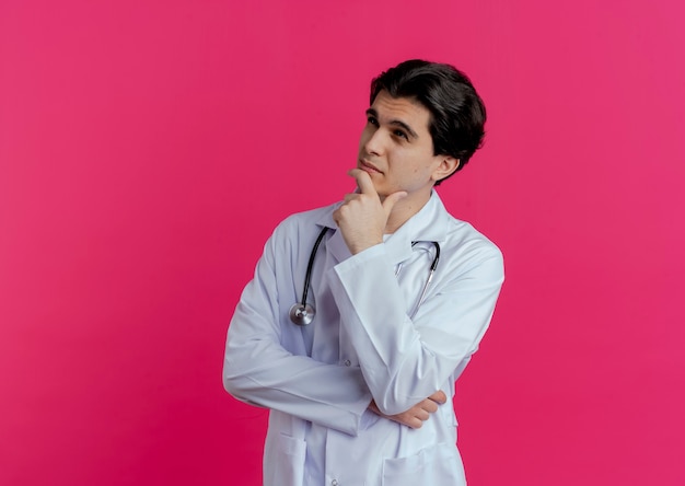 Nachdenklicher junger männlicher Arzt, der medizinisches Gewand und Stethoskop trägt, das Seite berührt, die Kinn berührt, lokalisiert auf rosa Wand mit Kopienraum