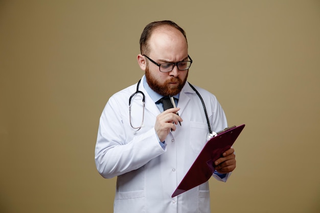 Nachdenklicher junger männlicher Arzt mit Brille, Laborkittel und Stethoskop um den Hals, der das Kinn mit Stift berührt und isoliert auf olivgrünem Hintergrund betrachtet