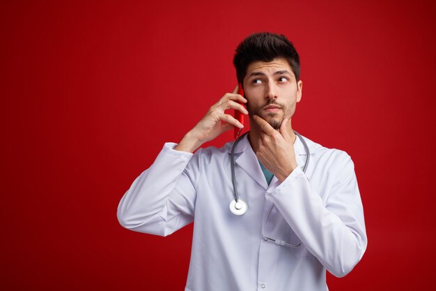 Nachdenklicher junger männlicher Arzt mit medizinischer Uniform und Stethoskop um den Hals, der die Hand am Kinn hält und am Telefon spricht und die Seite isoliert auf rotem Hintergrund betrachtet