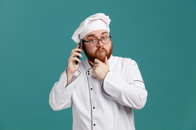 Nachdenklicher junger männlicher Koch mit einheitlicher Brille und Mütze, der die Hand am Kinn hält und in die Kamera schaut, die am Telefon isoliert auf blauem Hintergrund spricht