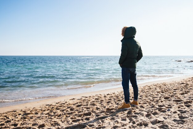 Nachdenklicher junger Mann, der das Meer, stehend auf dem Sand betrachtet. Rückansicht.