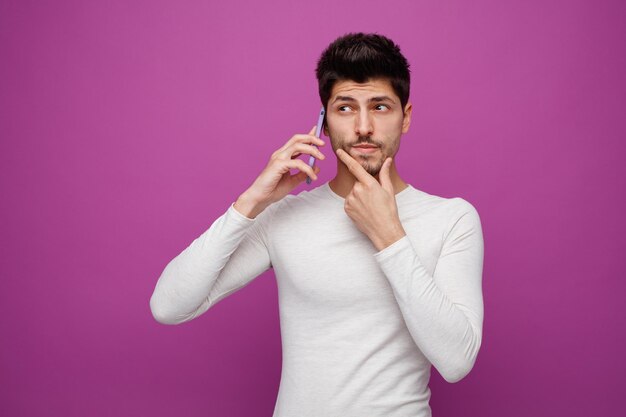 Nachdenklicher junger Mann, der die Hand am Kinn hält und zur Seite schaut, während er telefoniert, isoliert auf violettem Hintergrund