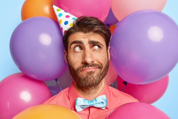 Nachdenklicher junger Mann, der durch bunte Luftballons des Geburtstages aufwirft