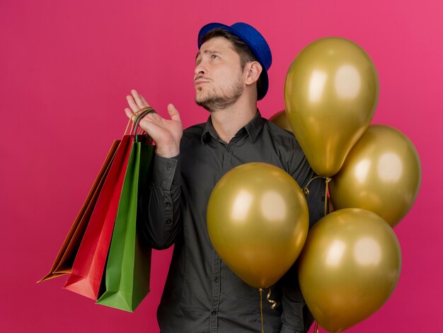 nachdenklicher junger Party-Typ, der blauen Hut hält, der Luftballons mit Geschenkbeuteln lokalisiert auf Rosa hält