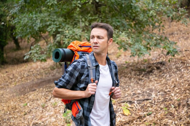 Nachdenklicher Mann, der im Wald wandert und Rucksack trägt. Männlicher Wanderer, der zur Seite schaut und im Wald geht. Kaukasischer männlicher Reisender draußen in der Natur. Tourismus-, Abenteuer- und Sommerferienkonzept