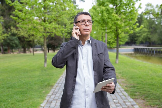 Nachdenklicher Mann, der Tablette verwendet und um Telefon im Park ersucht