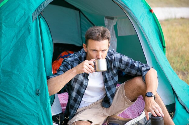 Nachdenklicher Mann, der Tee trinkt, im Zelt sitzt und wegschaut. Kaukasischer hübscher Reisender, der auf Rasen im Park kampiert und auf Natur entspannt. Backpacking Tourismus, Abenteuer und Sommerurlaub Konzept